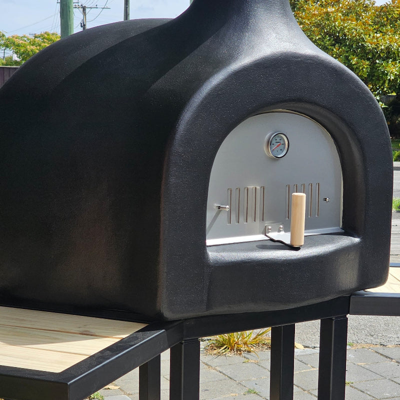 Black outdoor pizza oven with a thermometer on a stand outdoors.