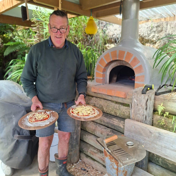 Man holding two pizzas in front of an outdoor brick oven