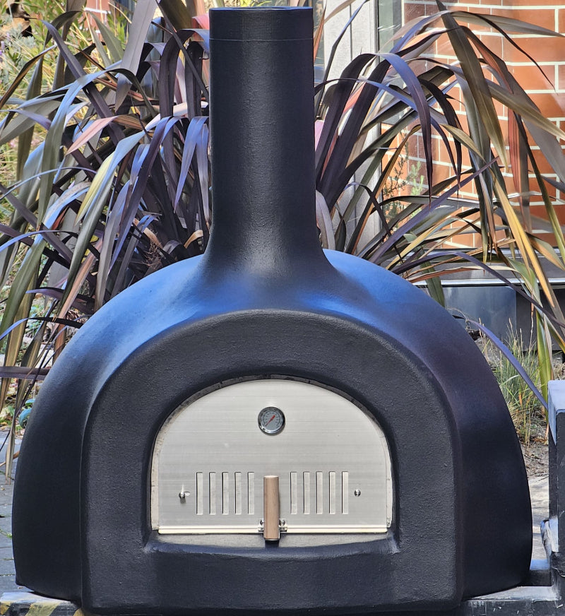 Black outdoor pizza oven with a brick building and plants in the background