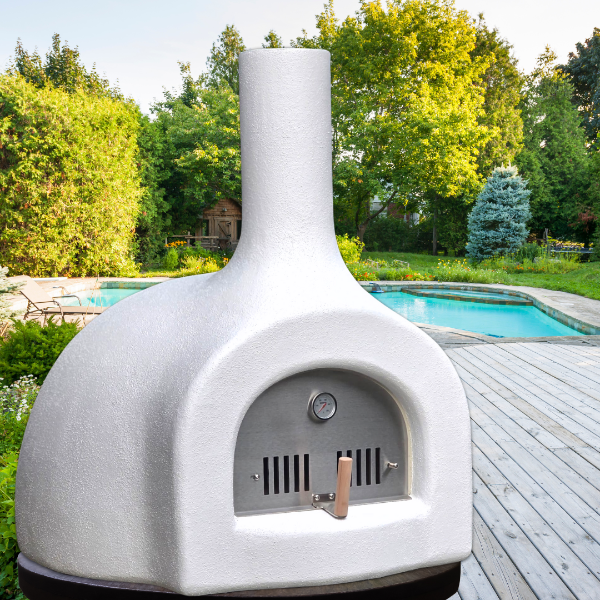 White outdoor pizza oven on a wooden deck with a pool and trees in the background