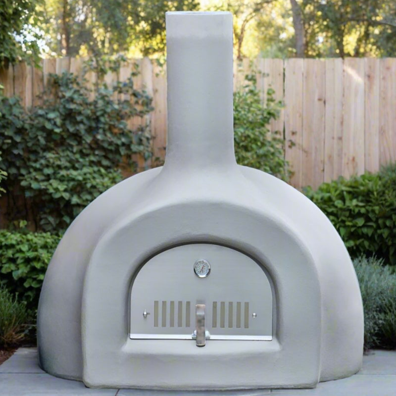 A grey outdoor pizza oven set in a garden with lush greenery and a wooden fence.